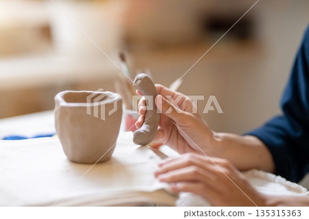 Close up potter artist holding adding handle to clay mug on working table in pottery class or studio Close up potter artist holding adding handle to clay mug on working table in pottery class or studio 135315363