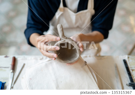 Pottery artist adding a handle to a clay mug over throwing wheel at working table in a class or studio. 135315367