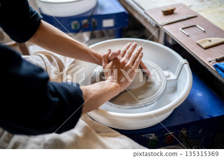 Close up of pottery artist's hands shaping clay body on spinning throwing wheel in a class or studio Close up of pottery artist's hands shaping clay body on spinning throwing wheel in a class or studio 135315369