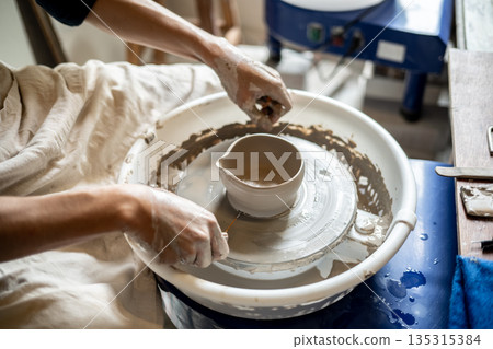 Close up pottery artist with a clay tea brewing bowl on spinning throwing wheel in class or studio Close up pottery artist with a clay tea brewing bowl on spinning throwing wheel in class or studio 135315384
