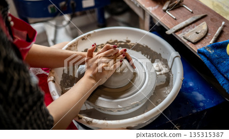 Woman pottery artist with painted nails molding a clay on spinning throwing wheel in class or studio Woman pottery artist with painted nails molding a clay on spinning throwing wheel in class or studio 135315387