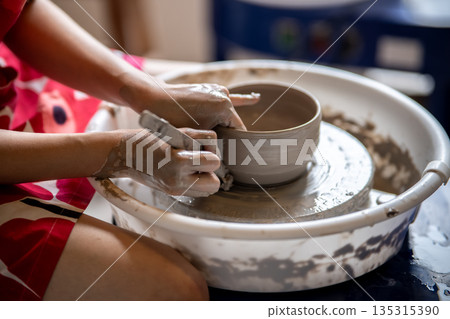 Woman pottery artist using tool scraping clay tea brewing bowl on throwing wheel in class or studio Woman pottery artist using tool scraping clay tea brewing bowl on throwing wheel in class or studio 135315390