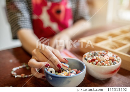 Painted nails woman artist threading ceramic beads making a bracelet or necklace in class or studio. 135315392