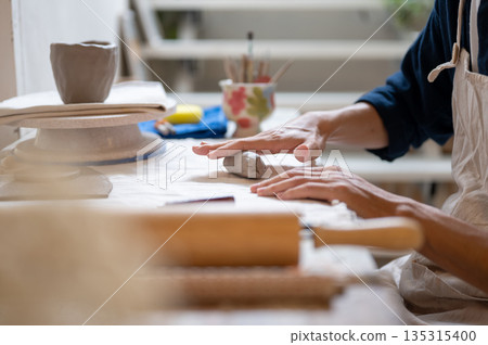 Close up of pottery artist's hand molding a clay mug's handle at working table in a class or studio. Close up of pottery artist's hand molding a clay mug's handle at working table in a class or studio. 135315400