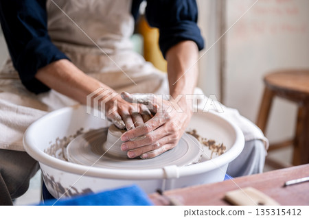 Close up of pottery artist's stained hands covering a clay on throwing wheel in a class or studio. Close up of pottery artist's stained hands covering a clay on throwing wheel in a class or studio. 135315412