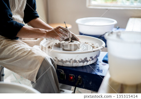 Close up of pottery artist using tool making a hole on clay on throwing wheel in class or studio. Close up of pottery artist using tool making a hole on clay on throwing wheel in class or studio. 135315414