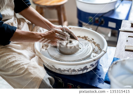 Close up of pottery artist using tool opening a hole on clay on throwing wheel in class or studio. Close up of pottery artist using tool opening a hole on clay on throwing wheel in class or studio. 135315415
