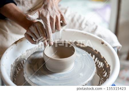 Close up pottery artist adding pour spout to clay brewing bowl on throwing wheel in class or studio. Close up pottery artist adding pour spout to clay brewing bowl on throwing wheel in class or studio. 135315423