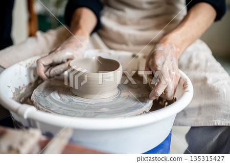 Close up potter using wire cutter on clay bowl for tea brewing on throwing wheel in class or studio. Close up potter using wire cutter on clay bowl for tea brewing on throwing wheel in class or studio. 135315427