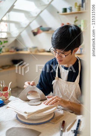 Young man potter artist taking out a circle cut clay on working table in a pottery class or studio. Young man potter artist taking out a circle cut clay on working table in a pottery class or studio. 135315506