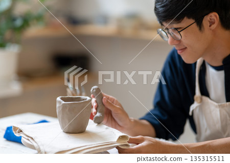 Potter artist man holding and adding handle to clay mug on working table in pottery class or studio 135315511