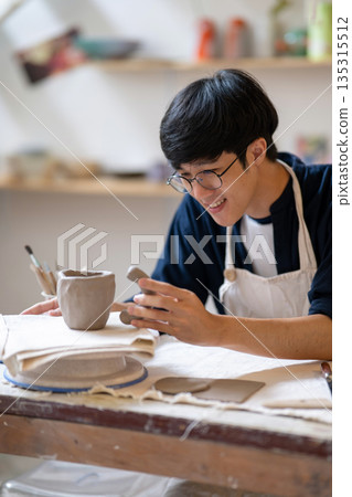 Potter artist holding and adding handle to clay mug cup on working table in a pottery class or studio 135315512