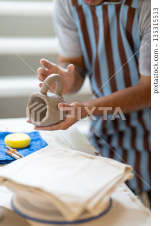 Potter artist holding and adding handle to a mug cup on working table in a pottery class or studio. 135315513