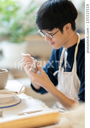 Pottery artist man using knife scraping on clay handle of a mug at working table in a class or studio. Pottery artist man using knife scraping on clay handle of a mug at working table in a class or studio. 135315519