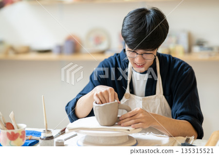 Young man pottery artist molding clay mug cup on throwing wheel at working table in class or studio Young man pottery artist molding clay mug cup on throwing wheel at working table in class or studio 135315521