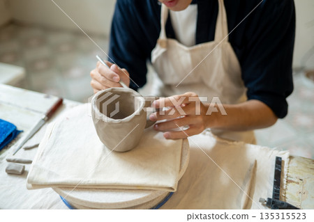 Potter artist using brush adding texture on clay mug at working table in a pottery class or studio 135315523