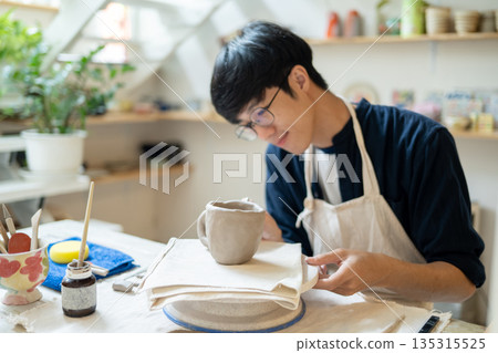 Young man potter artist looking closely at a clay mug at working table in a pottery class or studio. 135315525