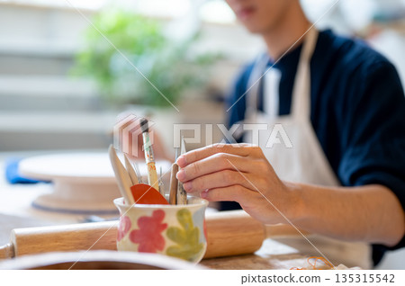 Young pottery artist taking a painting brush from ceramic cup on working table in a class or studio. 135315542