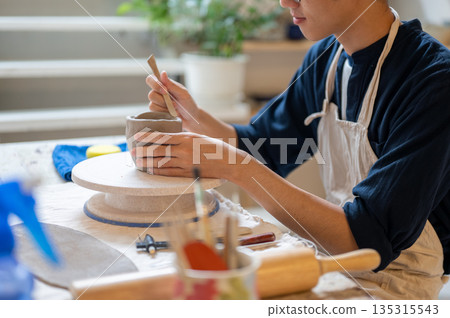 Young pottery artist using a throwing tool to make clay mug at working table in a class or studio. 135315543