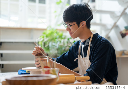 Glasses pottery artist using a throwing tool to make clay mug at working table in a class or studio 135315544