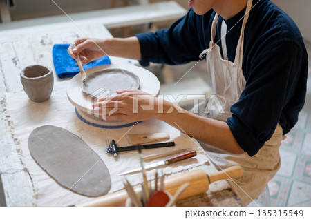 Pottery artist using modeling tool on clay slab on throwing wheel at work table in class or studio 135315549