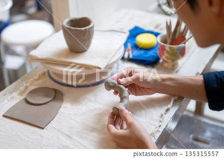 Close up of pottery artist's hand molding clay into a mug handle at working table in class or studio Close up of pottery artist's hand molding clay into a mug handle at working table in class or studio 135315557