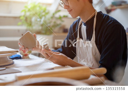 Young pottery artist holding and looking at clay mug with handle at working table in class or studio Young pottery artist holding and looking at clay mug with handle at working table in class or studio 135315558