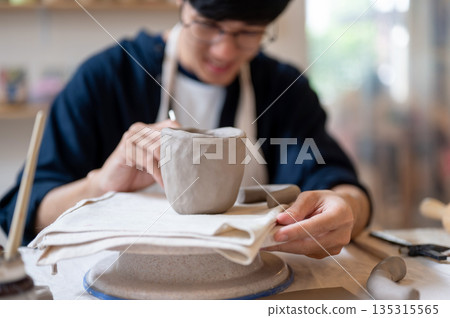 Pottery artist using tool adding texture to clay mug on a wheel at working table in class or studio Pottery artist using tool adding texture to clay mug on a wheel at working table in class or studio 135315565
