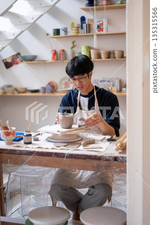 Pottery artist holding handle to a clay mug on throwing wheel at working table in a class or studio. 135315566