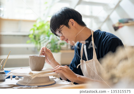 Pottery artist holding brush adding clay slip to glue mug handle at working table in a class or studio. Pottery artist holding brush adding clay slip to glue mug handle at working table in a class or studio. 135315575