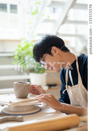 Pottery artist holding brush marking clay mug to glue a handle at working table in a class or studio 135315576