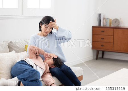 A mother takes her daughter's temperature by placing her hand on her forehead as she sleeps on her lap 135315619