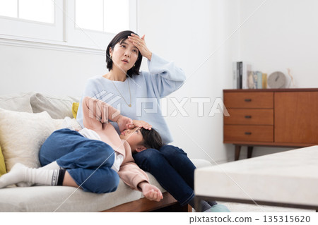 A mother takes her daughter's temperature by placing her hand on her forehead as she sleeps on her lap A mother takes her daughter's temperature by placing her hand on her forehead as she sleeps on her lap 135315620