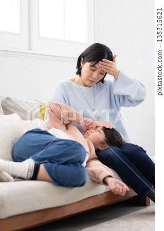A mother takes her daughter's temperature by placing her hand on her forehead as she sleeps on her lap 135315621