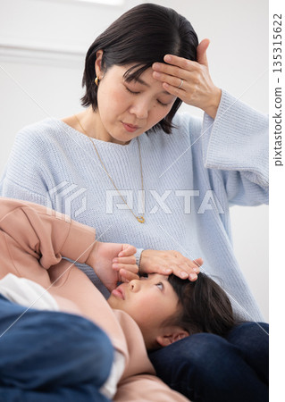 A mother takes her daughter's temperature by placing her hand on her forehead as she sleeps on her lap 135315622