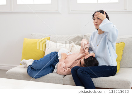 A mother takes her daughter's temperature by placing her hand on her forehead as she sleeps on her lap 135315623