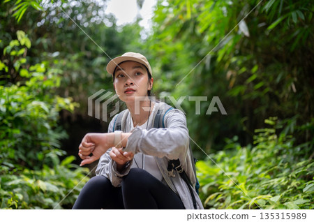 Woman backpacker sitting on ground looking away from watch while solo hiking or trekking in forest. 135315989