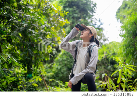 Backpack tourist woman holding binocular looking at wild bird on a solo hiking or trekking in forest 135315992