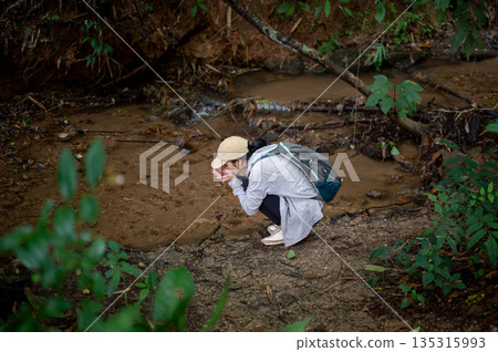 Woman tourist crouching near water stream to drink or wash her face during forest hiking or trekking 135315993