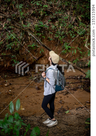 Woman tourist standing aside water stream looking up to waterfall during forest hiking or trekking. Woman tourist standing aside water stream looking up to waterfall during forest hiking or trekking. 135315994