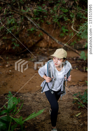 Woman tourist holding bag straps and walking up from a water stream during forest hiking or trekking 135315995