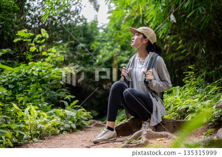 Woman backpacker holding bag strap sitting on ground tree root during solo forest hiking or trekking 135315999