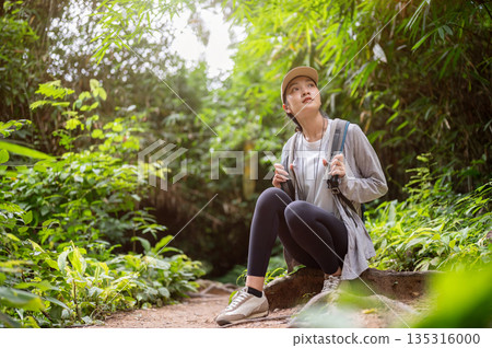 Woman backpacker holding bag strap sitting on ground tree root during solo forest hiking or trekking 135316000