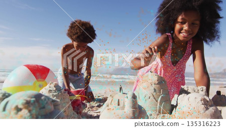 Siblings in pink dress and trunks building sandcastles on beach with red bucket, plastic shovel 135316223