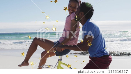 Smiling couple riding yellow bicycle along sandy beach in summer clothes, with golden star confetti Smiling couple riding yellow bicycle along sandy beach in summer clothes, with golden star confetti 135316227