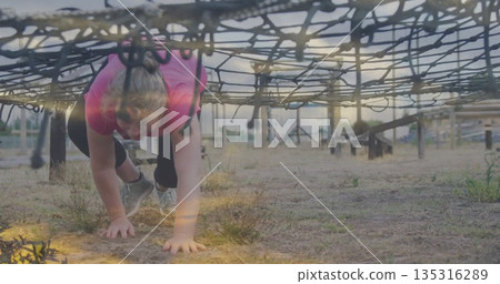 Athlete in pink sportswear crawling under cargo net in field, poles, grassy weeds, running shoes 135316289