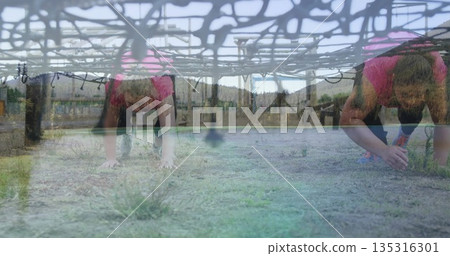 Crawling women wearing pink tops and leggings under rope net at obstacle course, with wooden posts 135316301