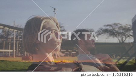 Rowing couple wearing sportswear pulling rowing machine handles in city park, with picnic tables 135316316