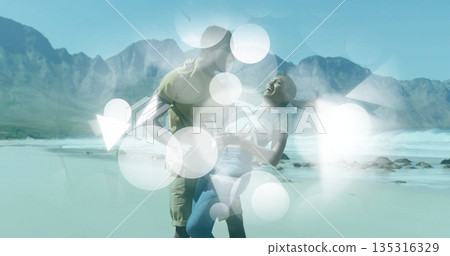 Romantic couple dipping and laughing on sunlit wet sand at beach shore, mountain peaks 135316329
