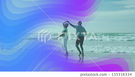 Dancing mature couple twirling barefoot at sandy beach, with rolling ocean waves and shallow water 135316334